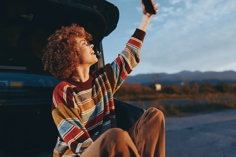 woman with curly hair in striped sweater sits by open car door, outdoor travel lifestyle, raising arm toward sunset, mountain landscape, warm evening light, casual mood, summer