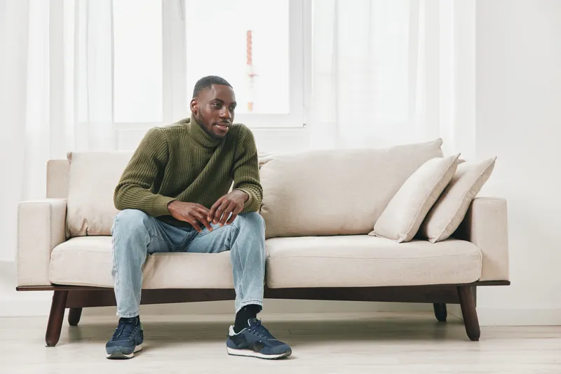 Young African man casually dressed in olive green sweater and jeans, sitting on a light colored couch in a modern interior, expressing contemplation and relaxation