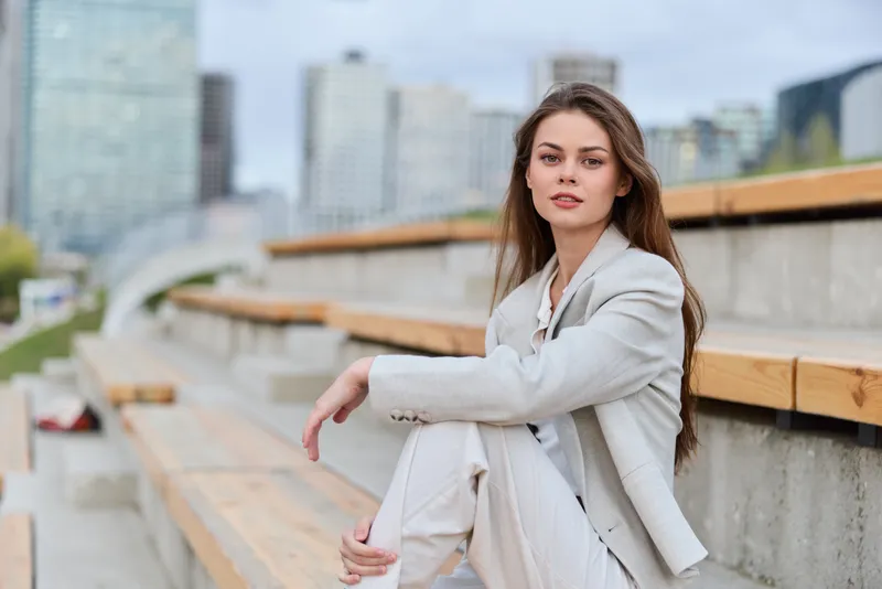 confident business woman sitting outdoors in stylish suit on urban bench with cityscape background, natural light, spring season, casual professional look