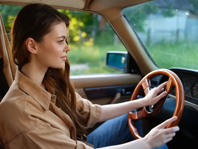 Young woman driving an old-fashioned car in a natural setting, showcasing confidence and tranquility, wearing a casual beige shirt, reflecting a vintage aesthetic.
