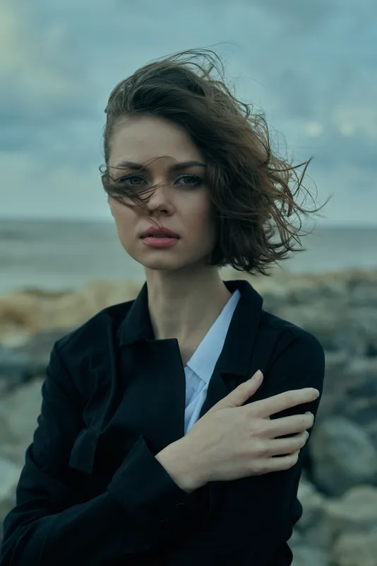 Woman standing on a windy coast in a dark outerwear jacket, hair blown by the wind, sunglasses on, thoughtful mood, rocky shoreline and ocean on the horizon, fashion and emotion in a coastal scene