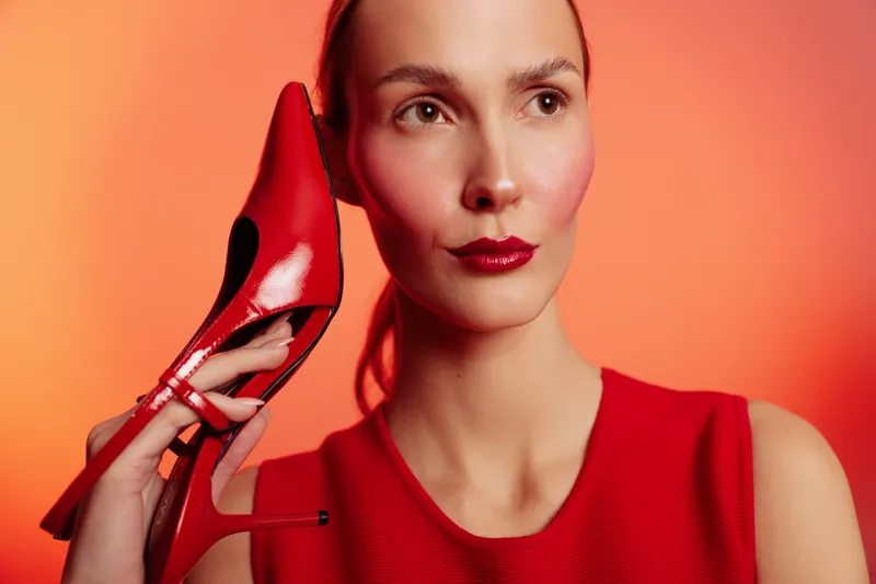 Elegant woman in red dress holds red high heel shoe near face against orange background. Fashion concept with stylish makeup and confident expression in studio lighting.