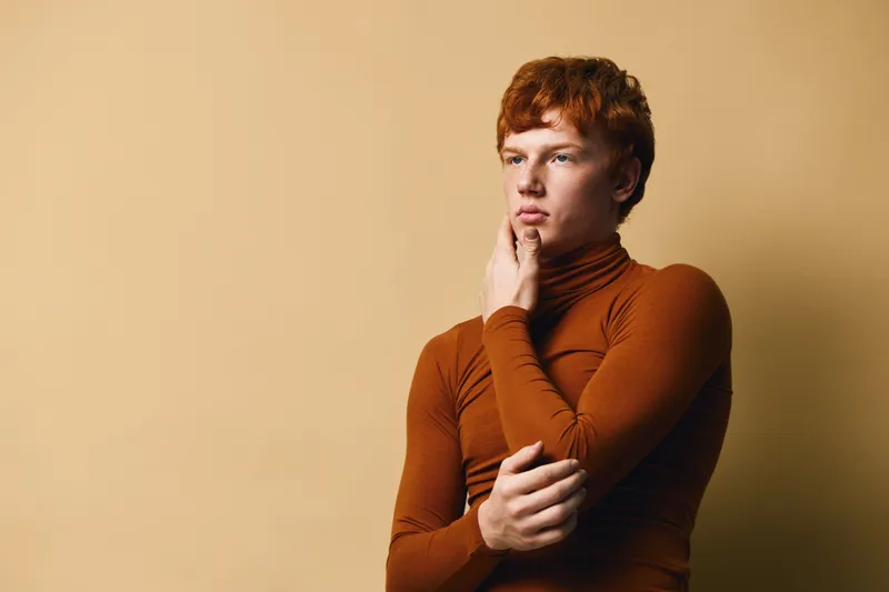 Young man with red hair and thoughtful expression wearing brown turtleneck posed against beige background with hand on chin and crossed arms.