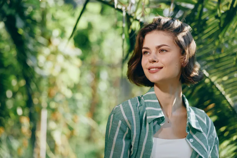 A young woman stands outdoors among lush greenery, smiling softly in a striped green shirt, captured in bright natural light.