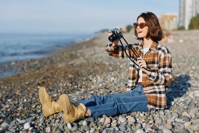 Woman photographer on a pebble beach, camera in hand, wearing a plaid shirt and jeans, enjoying outdoor photography by the sea with warm light and relaxed mood.