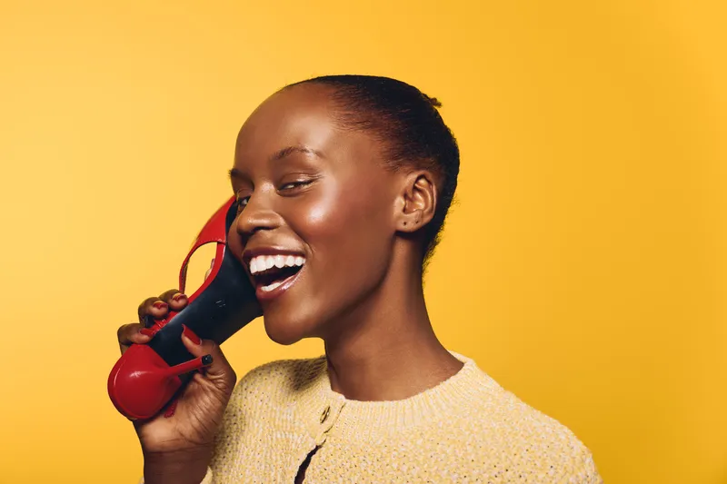 Happy African woman with dark skin laughing while holding a red vintage telephone handset on yellow background. Concept for communication, joy, and retro technology.