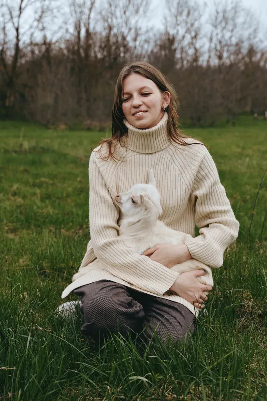 A calm woman in a chunky knit sweater sits on green grass, cradling a fluffy goat kid in a sunny field. Lifestyle scene portraying gentle bonding, farm life, and animal companionship.