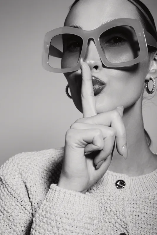 Stylish caucasian woman with fair skin and large sunglasses makes shushing gesture with finger on lips. Black and white portrait with focus on face and fashion concept.