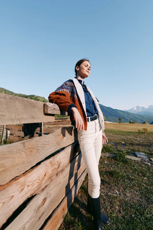 Woman standing and leaning on wooden fence in countryside field with mountains background, portrait style showing sweater, boots and casual fashion in scenic outdoor landscape.