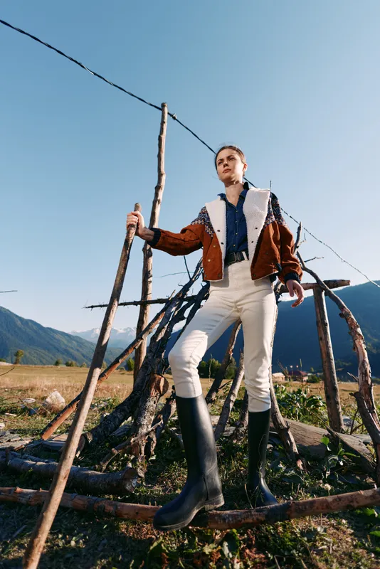 Hiker woman walking with stick and rubber boots wearing shearling jacket standing on wooden fence in mountains outdoors, portrait from low angle with clear sky and landscape.