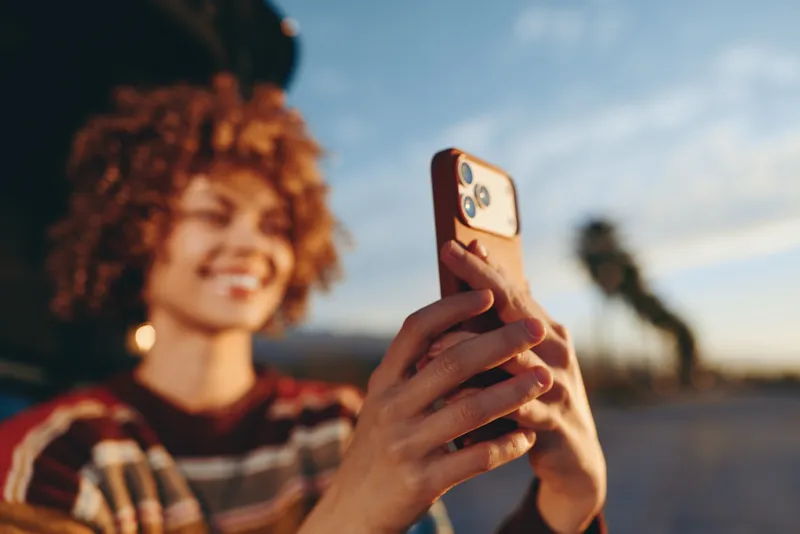 Woman lifestyle outdoors at sunset, holding a smartphone for a selfie, curly hair and friendly smile, casual sweater, warm evening light, blurred background, with bokeh and street walk