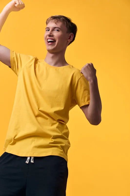 Celebration, happiness, victory, excitement on a youthful figure in a yellow tee, arms raised in triumph against a bright backdrop, conveying energy, confidence, and upbeat mood suitable for