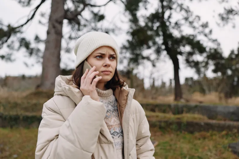 Woman outside in winter coat and beanie, talking on a phone with a pensive expression among trees in a chilly park setting.