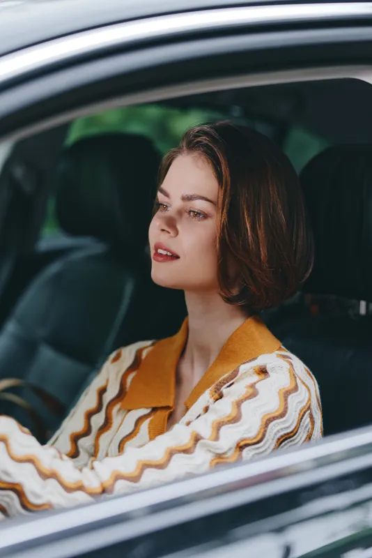Portrait of a woman driving a car, gazing ahead with a calm, confident expression. She wears a striped orange jacket, framed by the car window.