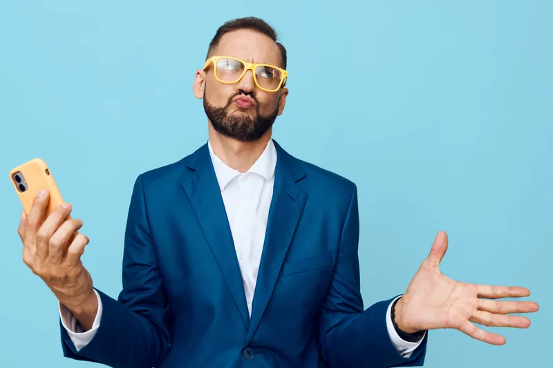 A confident business professional man stands against a solid blue background, wearing a navy suit and yellow glasses, holding a smartphone with a playful pose.