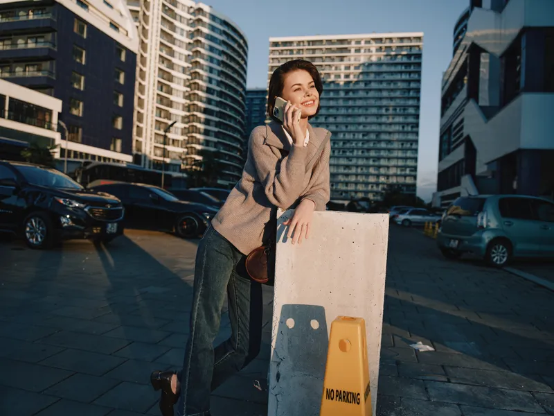 A cheerful woman stands outdoors in a modern urban setting, talking on a phone and smiling, leaning on a concrete barrier beside parked cars, conveying a friendly, confident worklife vibe.