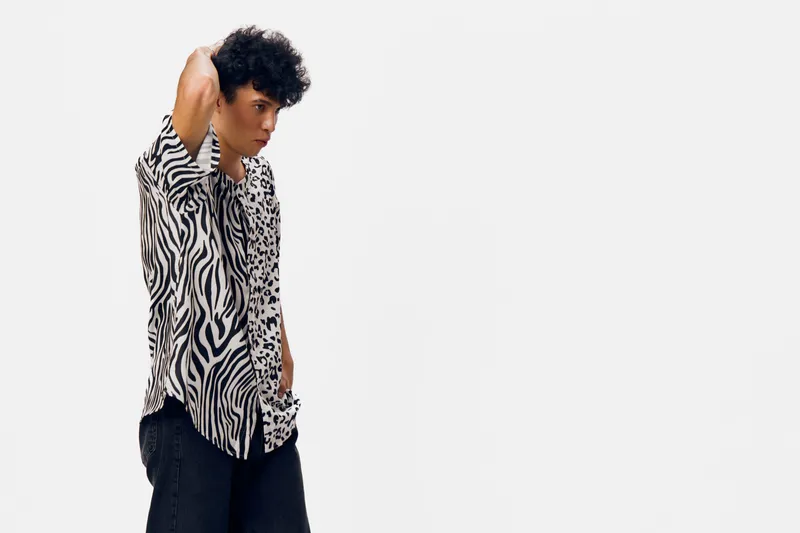 young man with curly hair wearing a black and white zebra print shirt and dark pants posing thoughtfully against a plain white background in a studio setting