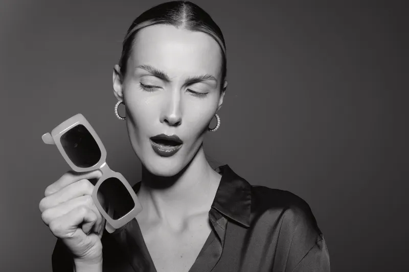 Young Caucasian woman with pale skin holds sunglasses near face, eyes closed, wearing dark blouse and hoop earrings. Studio portrait in black and white with elegant mood.