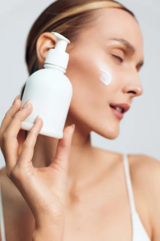 Young woman with blonde hair applies white cream on her cheek while holding a white pump bottle against a plain light background with closed eyes and relaxed expression.