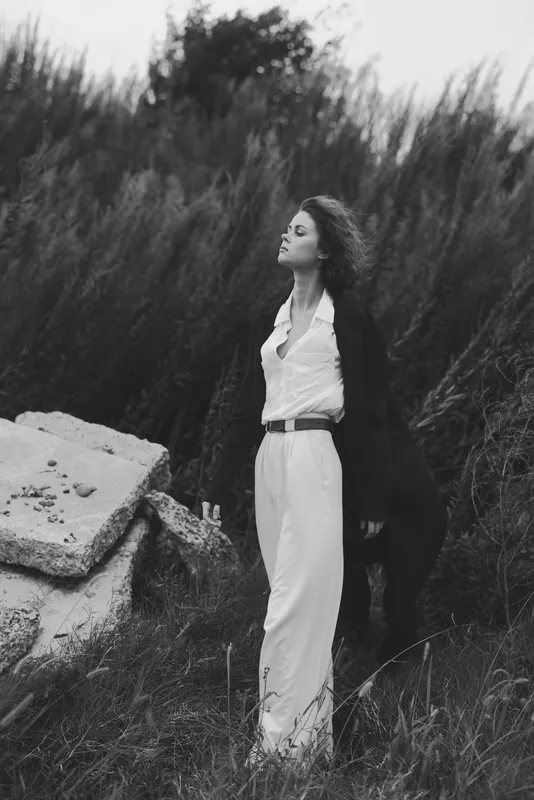 Woman in a white dress and a dark coat stands in tall grasses beside a stone structure, outdoors, wind in her hair, conveying elegant timeless beauty in a monochrome fashion shot.