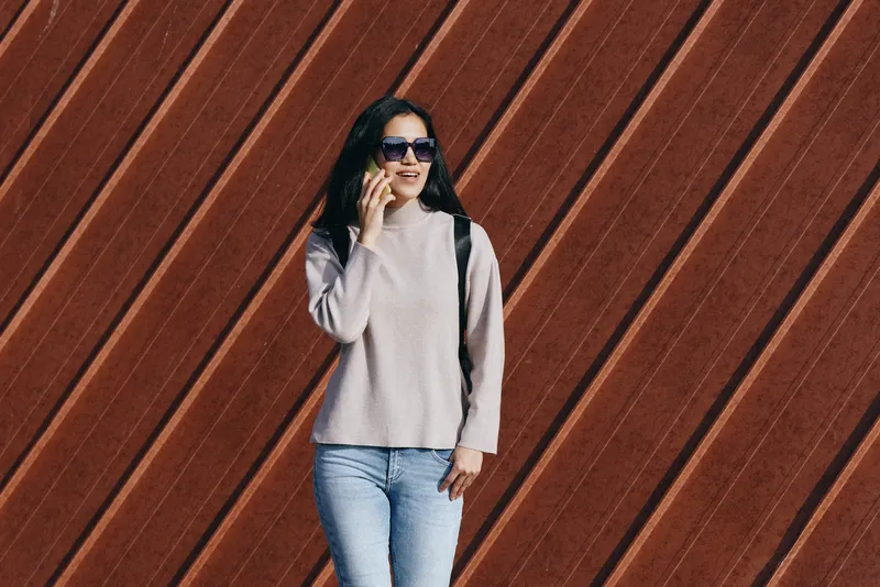 Woman in a light sweater, denim jeans and sunglasses stands with a backpack beside a diagonal wooden wall, capturing an outdoor urban lifestyle fashion moment