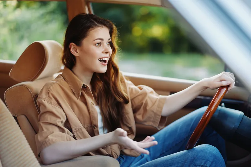 Happy young woman enjoying a joyride in a car, wearing casual clothing and expressing excitement in a green outdoor setting.