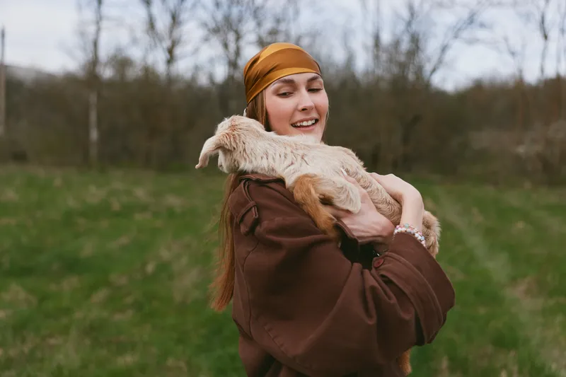 Joyful woman in a brown coat hugs a fluffy goat kid in an open field, showcasing rural lifestyle and tender bond, bright natural light, casual outdoor moment, serene countryside mood.
