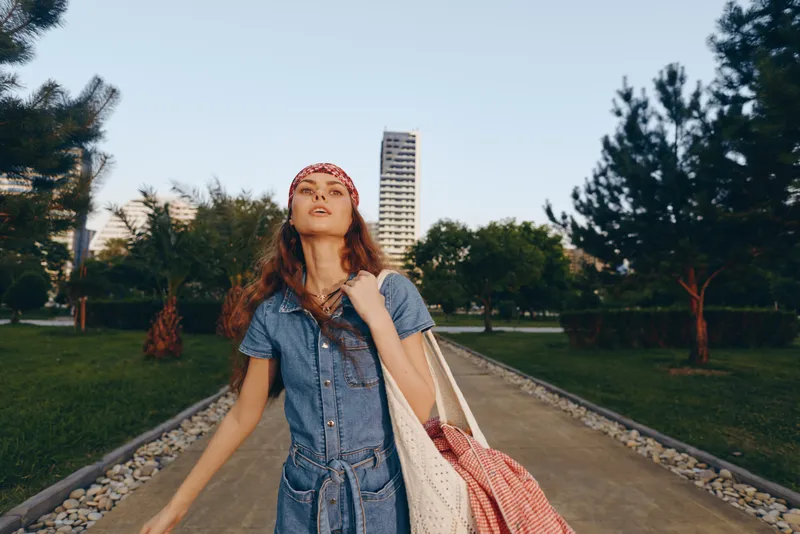 lifestyle woman in modern boho-western denim street style poses confidently outdoors with film color effect, wearing a headband and carrying a tote bag in a sunny park setting