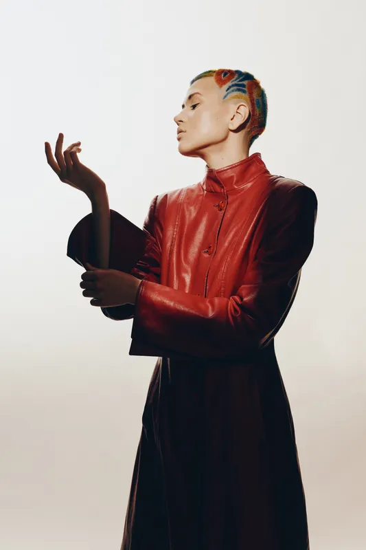 Woman with colorful braided hair wearing a red leather jacket poses gracefully against a plain light background, showcasing modern fashion and bold style choices.