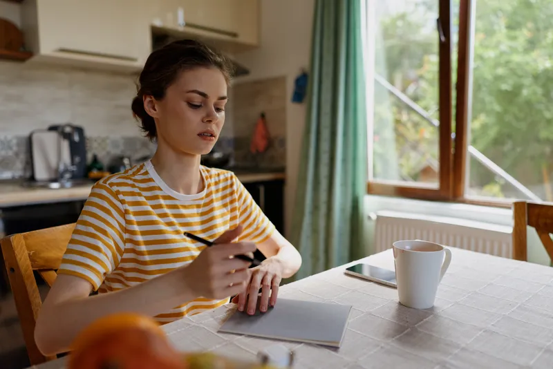 Woman at home writes on a notepad, casual striped shirt, sits at table by window, natural light, coffee mug nearby, focused moment of everyday creativity in a cozy kitchen.