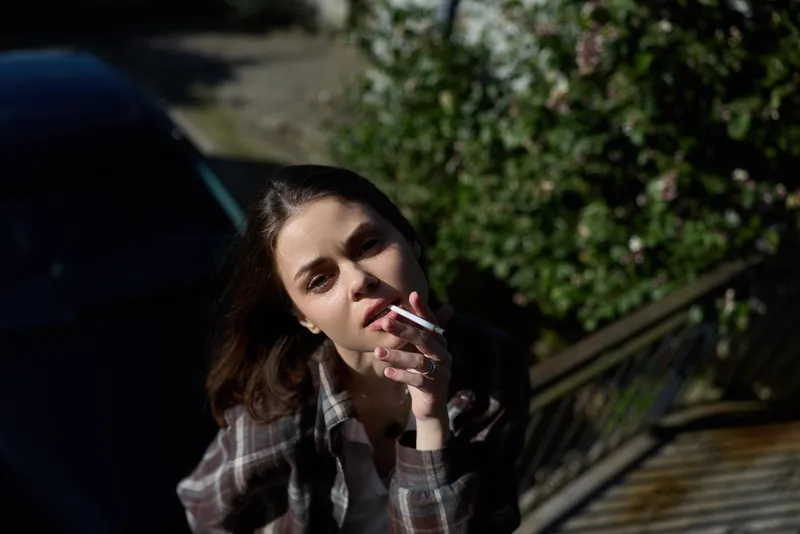 A woman outdoors in daylight calmly smokes a cigarette, wearing a casual plaid shirt, contemplative expression. A candid lifestyle moment