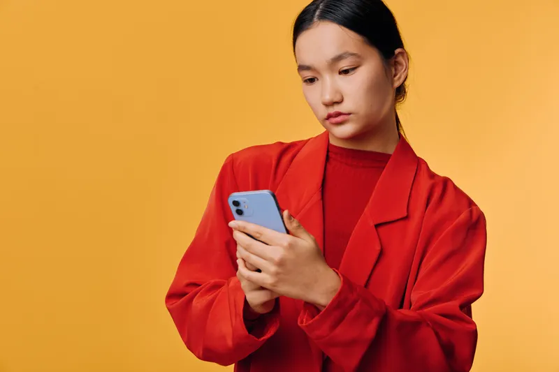 Woman in red jacket holding smartphone with focused expression on yellow background. Female using mobile device for texting or browsing with casual look and dark hair tied back.