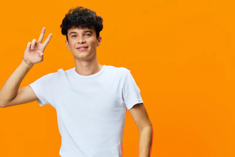 Young man in white t-shirt showing peace sign with smile, curly hair, standing against bright orange background expressing positivity and youthfulness.