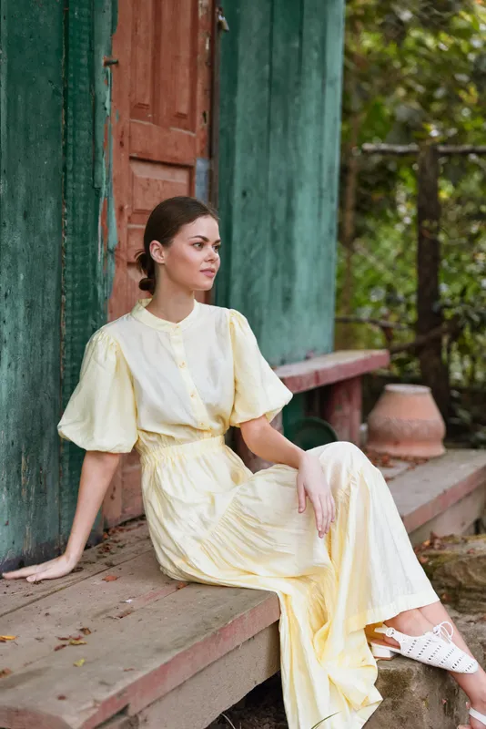 A woman in a pale yellow dress sits on steps beside a green door in an outdoor setting, radiating calm, grace, and relaxed fashion.
