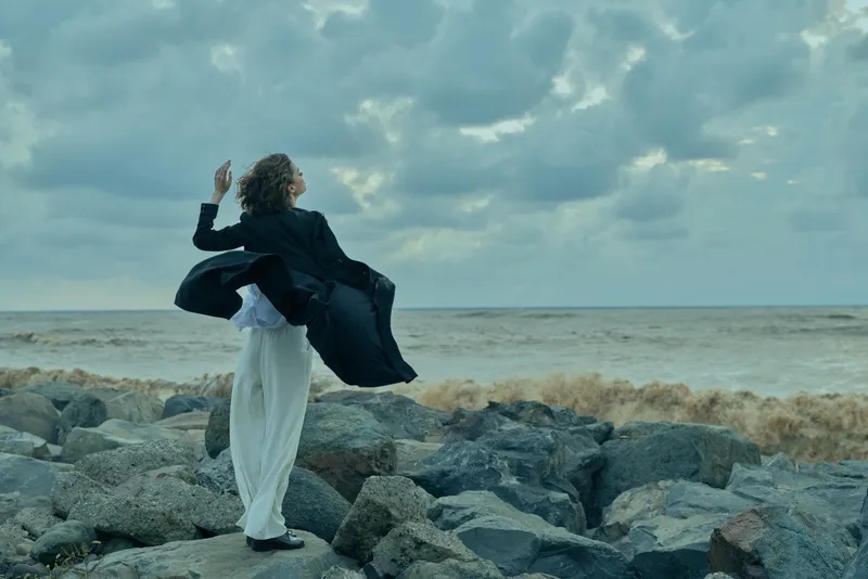 Windy coastline scene with dramatic sea and rocky shore, a solitary figure in a dark coat and white pants stands on stones as wind swirls fabric beneath a stormy sky