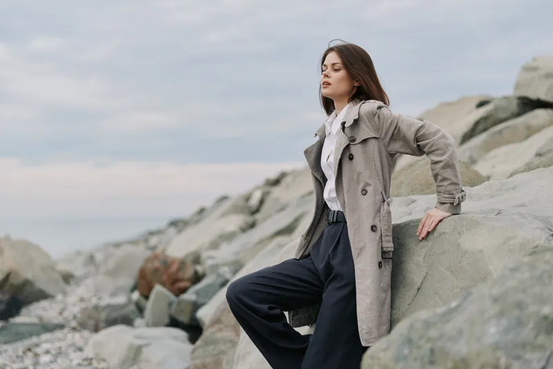 Young woman in a long coat leans on rocks at the beach under a cloudy sky. She looks thoughtfully into the distance, wearing dark pants and a white blouse. Concept for fashion and nature.