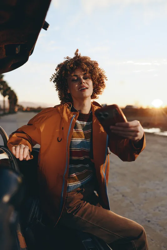 Woman lifestyle, curly hair, orange jacket, outdoors sunset scene near car, travel on road, young woman sitting outdoors, holding phone, warm golden sunlight, casual portrait