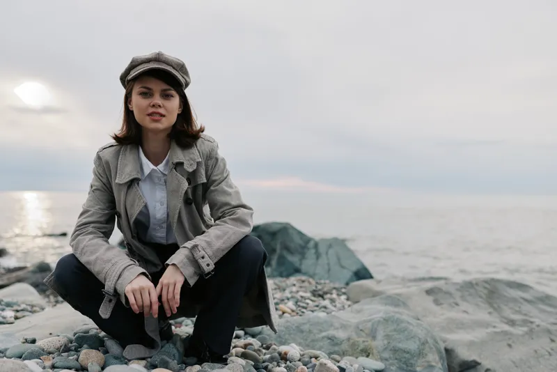 Young woman in stylish coat and cap squats on rocky beach near calm sea under cloudy sky. Concept of autumn fashion, outdoor lifestyle, and natural beauty by the water.