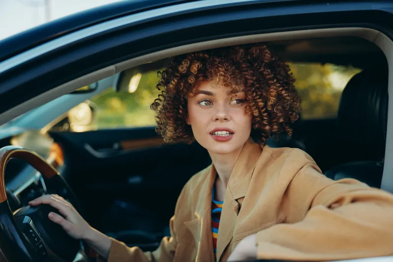 Woman lifestyle shot inside a car, retro film color, curly hair, blazer, looking ahead with curiosity, candid drive scene in urban setting, the composition highlights a stylish, independent mood and