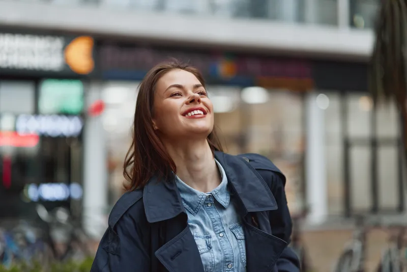 Smiling woman with long brown hair wearing a denim shirt and dark jacket standing outdoors in an urban environment during daytime.