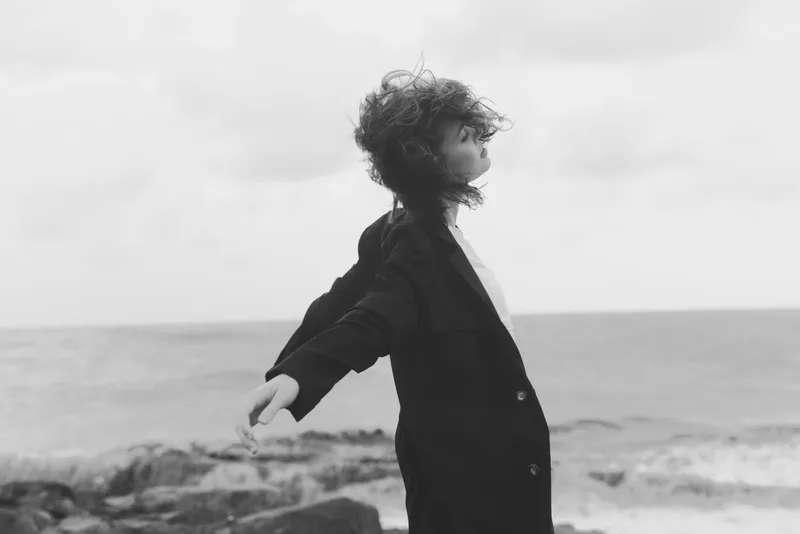 Child stands on windy shore with arms outstretched, coat billowing, hair swept by ocean breeze, black and white scene capturing freedom, solitude, and momentary escape by the sea, minimal composition
