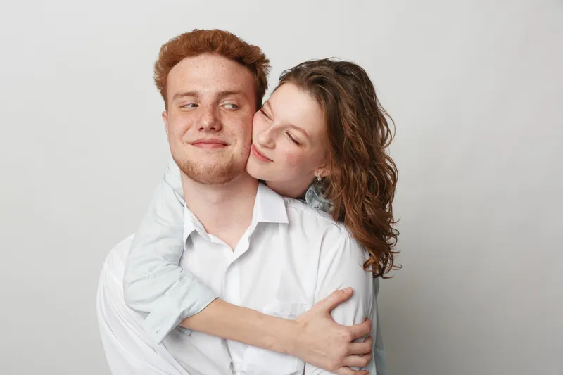 Young couple in casual clothes embraces tenderly against a plain background. The woman hugs the man from behind, both showing affection and warmth. Concept of love and intimacy.