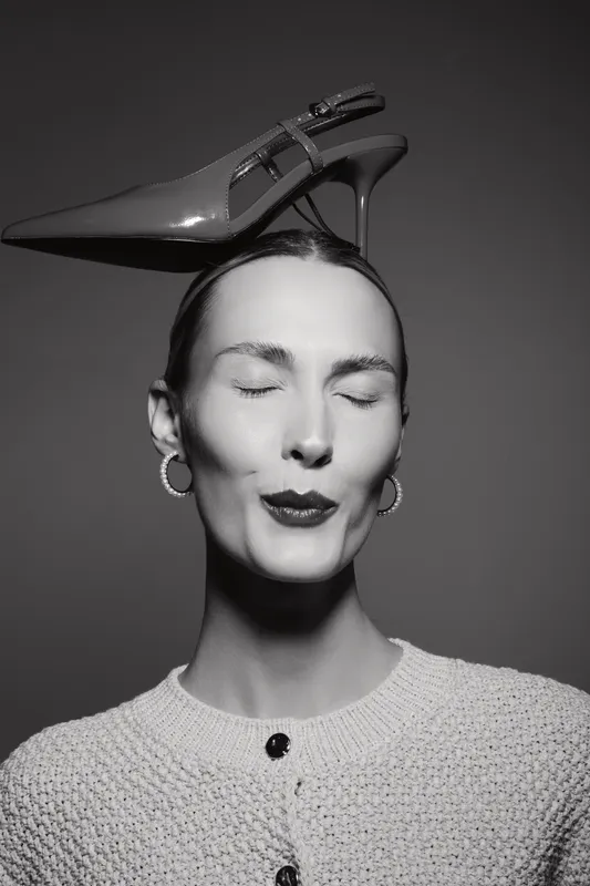 Portrait of a light skinned Caucasian woman with closed eyes balancing a high heel shoe on her head. Black and white studio lighting highlights fashion and creative beauty concept.