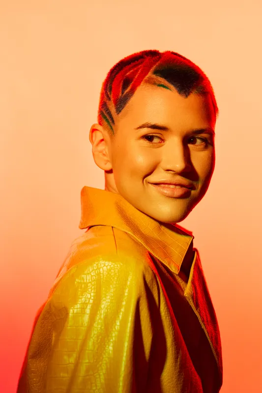 young woman with red hair and stylish haircut wearing yellow leather jacket smiling confidently against warm orange background in studio lighting