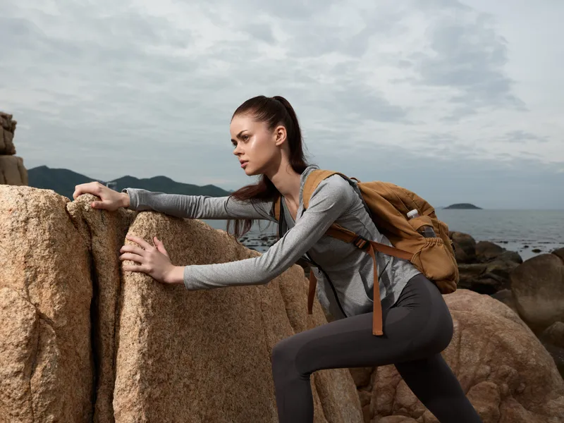 Young woman in activewear climbing rocks by the sea, embodying determination and adventure, with a brown backpack, against a cloudy coastal backdrop