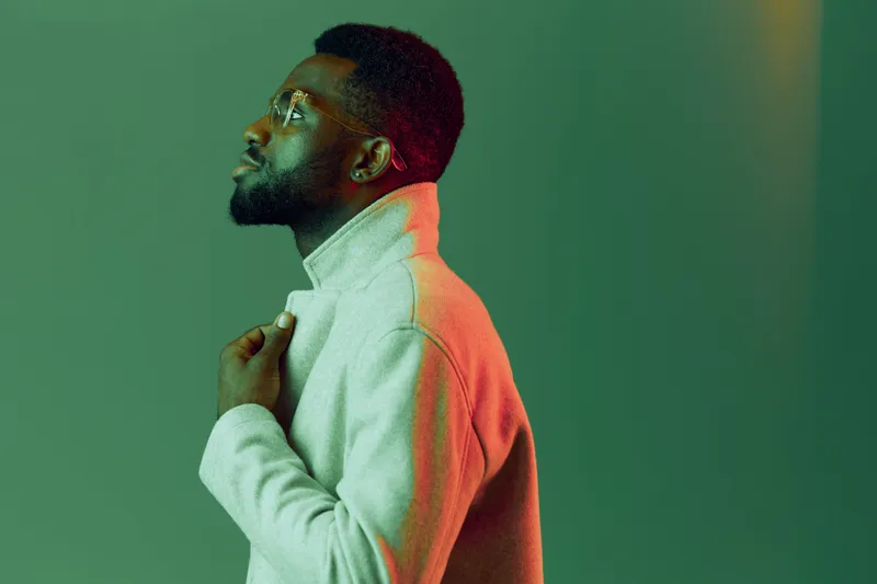 Stylish young man in a high neck sweater against a vibrant green background, expressing contemplation and confidence in a modern setting