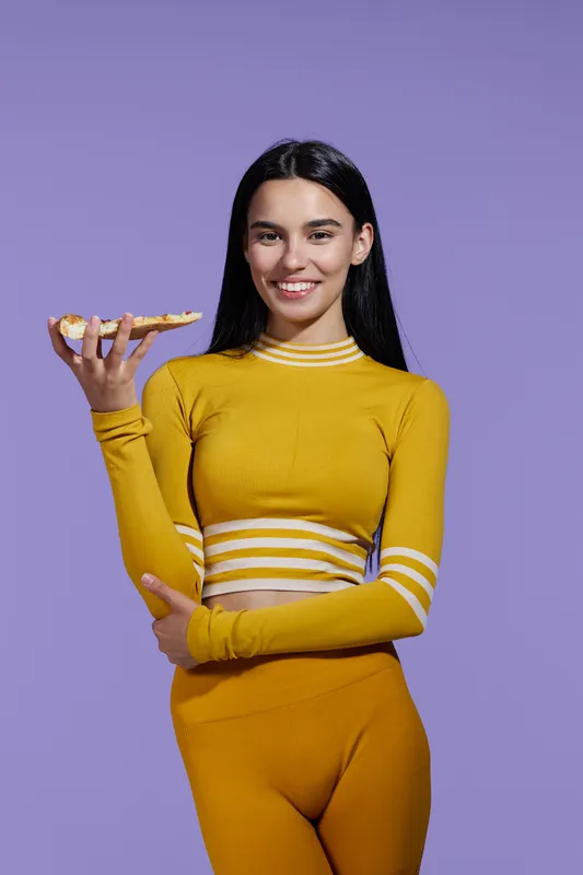 Sport fitness woman in yellow activewear smiling while holding a slice of pizza against purple background, showcasing healthy lifestyle and balance.