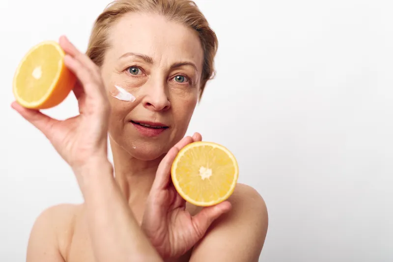 Mature woman holding two halves of an orange with facial cream on her cheek, showing natural skincare and healthy lifestyle concept with fresh citrus fruit and clean white background.