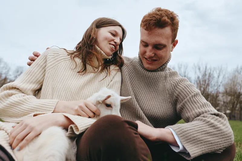 Smiling couple outdoors with a playful goat kid, sharing a cozy moment on a grassy field. Casual sweaters and soft natural light convey wellness, connection, and rural charm.
