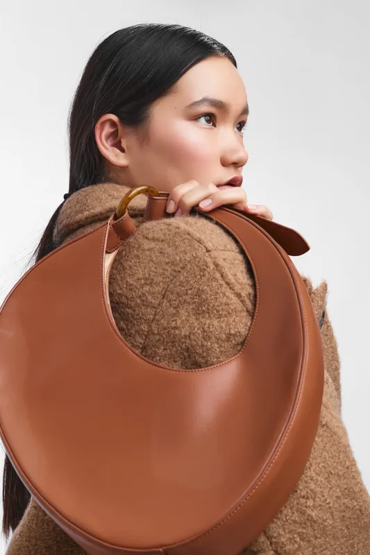 Asian woman presents a thoughtful pose as she rests a large round brown bag on her shoulder, creating a minimalist fashion moment in a studio setting.